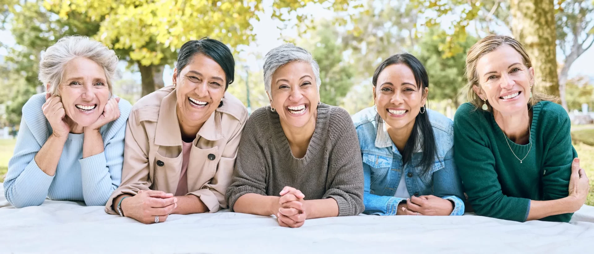 Five middle-aged women leaning forward together outdoors in a park, smiling warmly at the camera with trees and greenery in the background. Wellness - Menopause - Metabolic Health Consultations